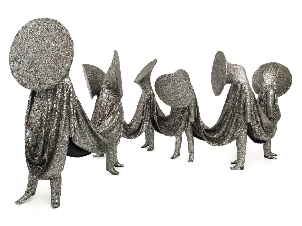 Seven silver/glittery "soundsuit" sculptures by Nick Cave standing in a line connected by what looks like draped fabric 