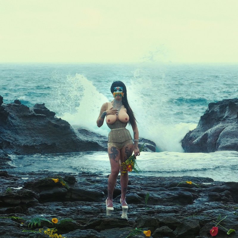 A person standing in front of a beach with a black wig, facepaint, breast forms, a corset and high heels, and flowers are scattered along the rocks