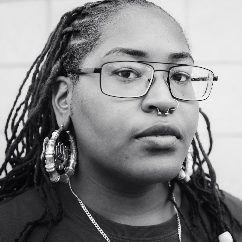 A closeup black and white photo of a black woman with glasses looking at the camera with a stoic expression
