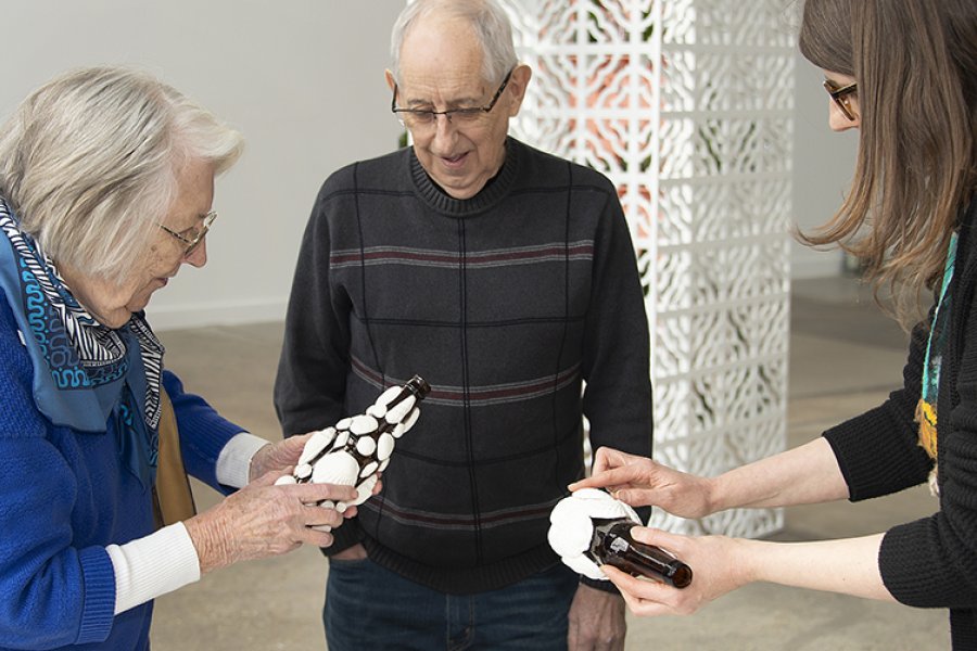 An older woman and a younger woman hold glass bottles with clay seashells glued to them while an older man looks on