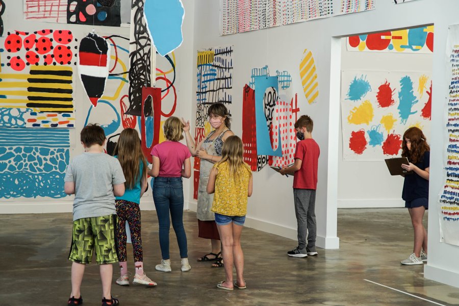 A woman and several kids standing in an exhibition space with colorful artworks that are predominantly red, yellow, and blue