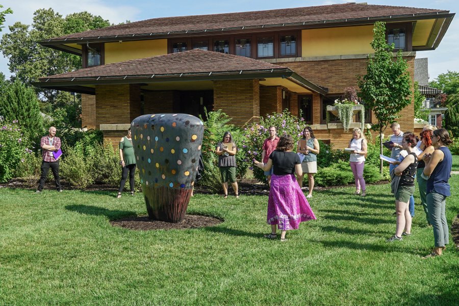 Adults standing around a large ceramic sculpture on a green lawn in front of a brown brick building