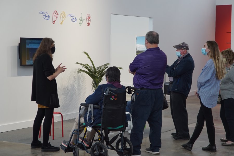 An educator gives a tour in front of an exhibition display with a television and sign language prints to a group of two men, two women, and one woman in a wheelchair. 