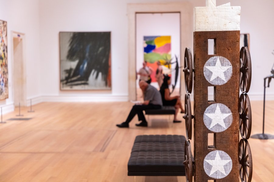 In the foreground is a wooden sculpture, behind which seated people sit amidst a brightly lit gallery