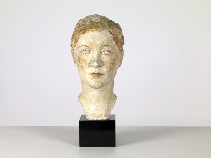 This realistic, but minimally detailed sculptural head of a woman is nearly life-sized and sits on a black, square base. Her short neck ending in a slight curve to indicate the shoulder line. Textured plaster and a palette of ivory, peach, ochre, and dark amber enrich feminine features, such as rounded lips and almond-shaped eyes accentuated by dark contours and pale pupils. Slicked-back, bobbed auburn hair and flushed, freckled cheeks, suggest that she is a modern woman of her time.