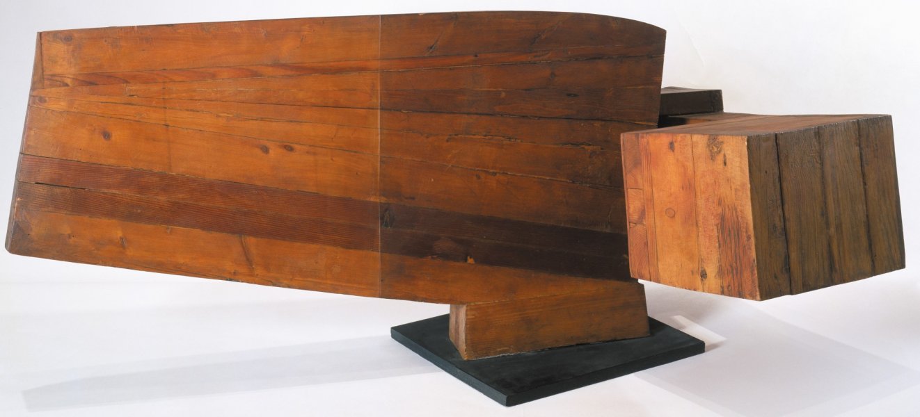 This horizontally expansive, wooden sculpture sits on a narrow base atop a small flat black square. Multiple horizontal and vertical layers of wood are joined and sanded smooth, revealing different colors and knots of the grain. A low horizonal rectangular shape juts far out on the left side, and gradually getting narrower. A cube-like structure with vertical layers protrudes out from the center of the sculpture and to the right.