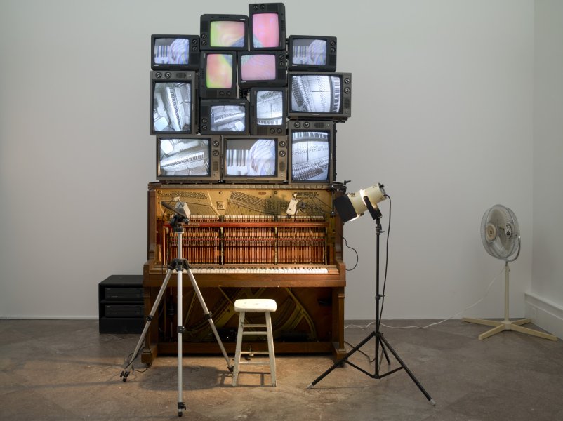 At the heart of this sculpture are thirteen boxy television sets of different sizes stacked atop of an upright player piano. Some screens livestream the moving keys as recorded by a video camera on a tripod set up in front of the piano while others show prerecorded footage of hands playing a piano. A white stool and a small spotlight on a tripod illuminating the keyboard are also set up in front of the piano.