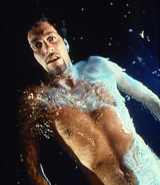 The first of three stills shows a nude man floating on his back in dark blue water, visible from the waist up. Light shines on his body from above, highlighting his face and part of his torso.