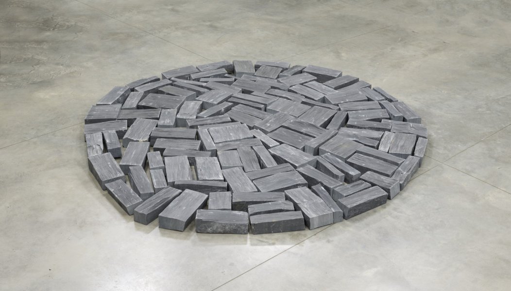 A number of rectangular bricks of natural slate of varying dimensions are packed together to form a nearly perfect circle. In this image, you are looking down at the work as installed on a light colored parquet floor from a shallow angle.