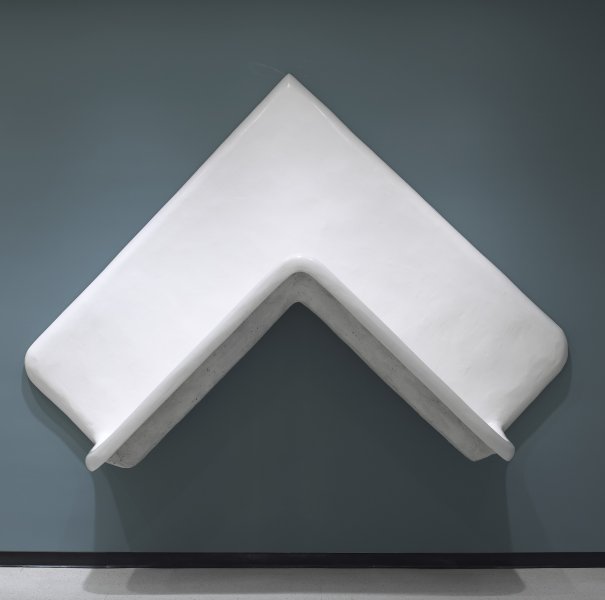 A large white plaster sculpture that takes the form of a thick, inverted V hangs from a blue-gray wall. The apex of the V comes to a sharp point whereas the left and right corners are curved.