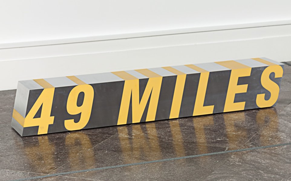 The phrase “49 MILES” in yellow block letters, tilted slightly as if set in italics, fills the polished aluminum surface of this sculpture facing the viewer. Additional stripes of yellow extend from where these numbers and letters touch the left and top edges of the sculpture’s front surface across its left and top surfaces. Overall, the sculpture takes the form of a rectangular cube whose depth and width are equal to one another and considerably smaller than its length. It is shown installed on the floor.