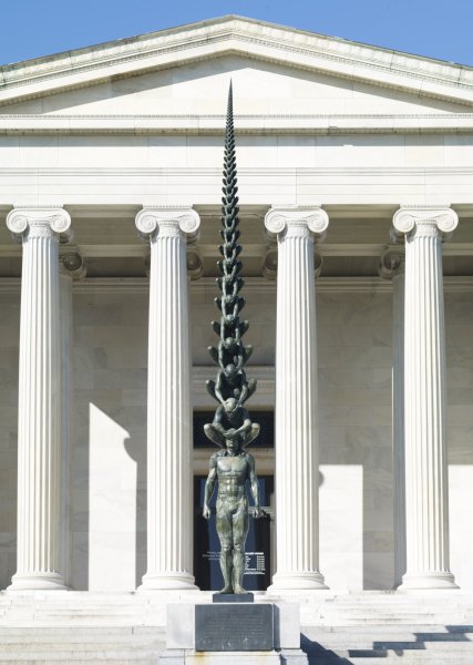 Against the white marbled columned exterior of the museum, a life-size muscular male nude, his arms hanging down on either side of his body, stands upright on a metal base. Sitting on his shoulders are a series of identical male nudes squatting one on top of the another, each one smaller than the one below. Each figure holds his hands over the eyes of the figure beneath him.