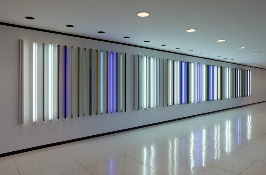 An installation of fluorescent light tubes mounted vertically with slight spaces in between extends along the white wall of a long hallway. The tubes glow in an array of colors, from bright white to numerous shades of blue, green, and brown. Their light is reflected in the hallway’s white tiled floor.