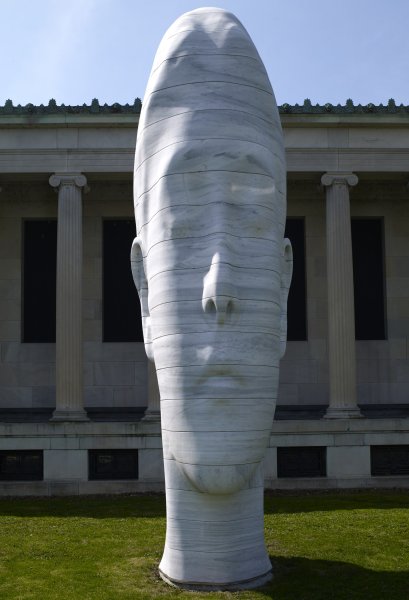The highly elongated head of a young woman with her eyes closed in layered slabs of white marble sits on a patch of grass in front of a portion of the museum’s columned façade.