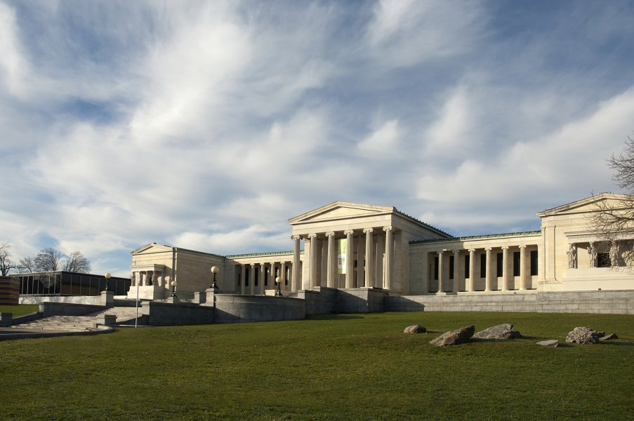 At bottom right of this color photograph, eight boulders are partially embedded in a circle in the lawn behind the museum. Each boulder differs slightly in size, texture, and shade of greyish brown. Bright sunlight enters the image from the top left, and the boulders cast long shadows on the surrounding grass. The museum’s neoclassical building and black glass addition span across the center of the image below a sky dotted with wispy clouds.