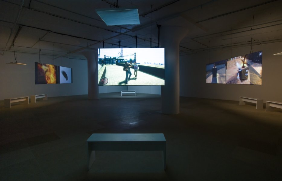 Five large digital screens hang from the ceiling of a darkened room. Two are hung side by side toward the left and rear of the room; two more are hung side by side toward the right and rear of the room; and a single screen hangs in the center foreground. All five screens feature imagery sourced from contemporary video games.