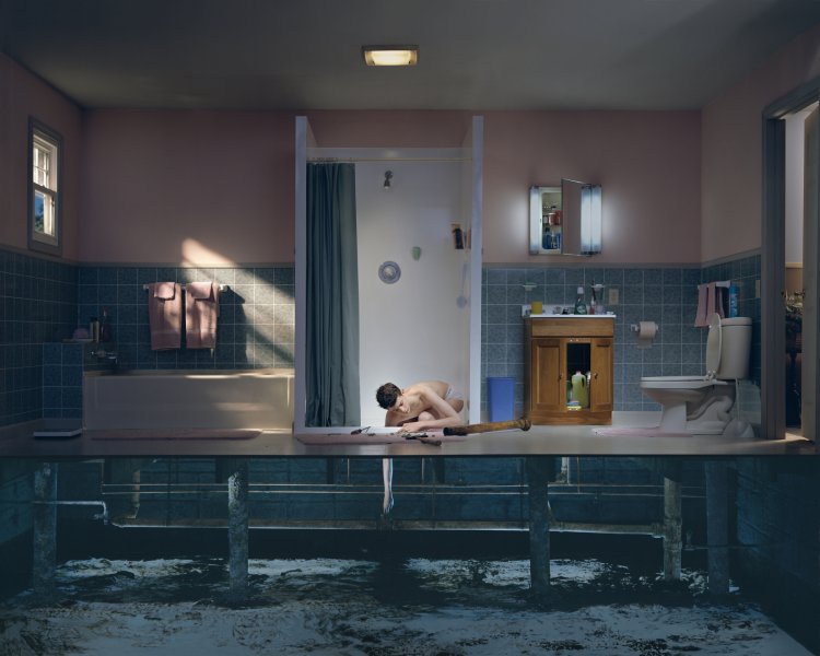 This large-scale color photograph captures a mysterious scene staged in a domestic bathroom, which is presented in cross-section. In the center of the frame, a white man, wearing only briefs, is bent over on the floor of a shower stall. His right arm reaches through the drain and is visible in the dark crawlspace below floor level where a pipe would normally be connected. The dim light filtering through a small window on the left evokes a sense of twilight.