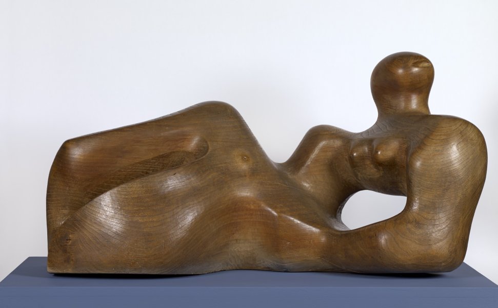 This wooden sculpture could abstractly resemble a reclining female body. The dark brown wood is polished to reveal the wood grain and knots throughout the form. The figure’s legs seem to be gently bent, and she appears to be resting on her elbows, with her head propped upright. The artist left the torso as an empty space, allowing viewers to see through the sculpture.