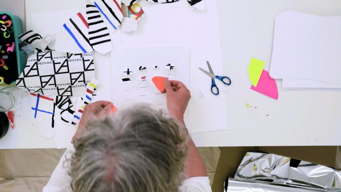 A bird's eye view of a man with gray hair placing a scrap of paper on a paper collage on the table in front of him