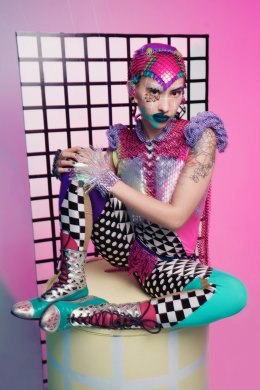 sky sits on a yellow pedestal with their feet up and their hands on their right knee. they are wearing colorful spandex suit that goes from their feet to the top of their head. it is turquoise, black and white checked/diamond/polkadots, and has shiny pink and silver scales with purple shoulder ruffles. they have olive skin and are wearing dark blue lipstick as they stare into the camera lens.