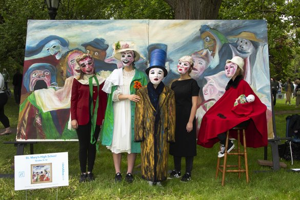 High school students wearing papier mache masks and dressed in elaborate historical dresses and suits stand in front of a painted background. The student on the right wears a red cape, sits on a stool, and holds a baby doll.
