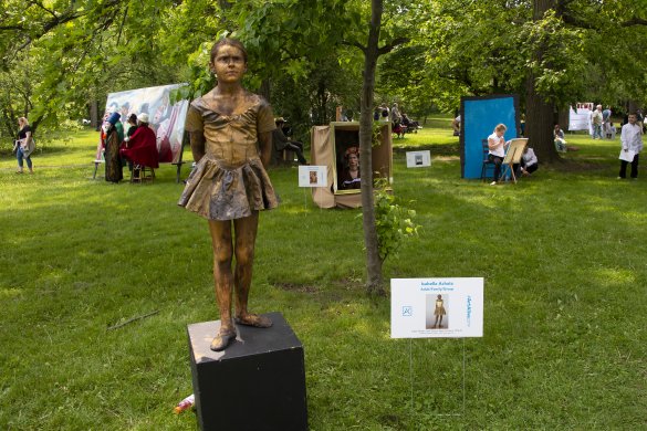 A girl dressed as a ballerina painted gold and standing on a pedestal outside on green grass