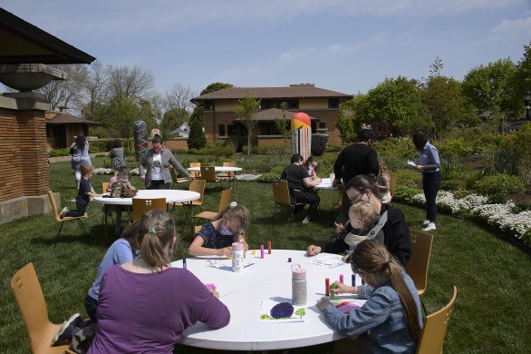In the foreground, people sitting at a table making art and, in the background, people looking at large ceramic sculptures on a green lawn in front of a brown brick house