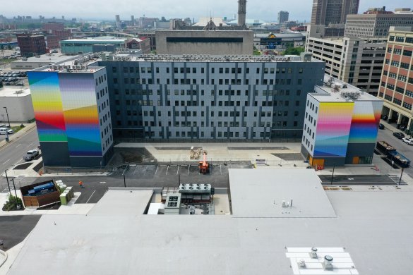 An elevated photograph of Josef Kristofoletti's Golden Hour, a mural that covers four faces of the building at 201 Ellicott Street in bands of bright color. In the background is the cityscape of Buffalo.