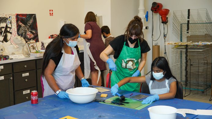 A woman in a green apron showing a teen girl how to use a roller to put paint on her print block