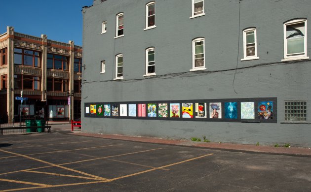 An exterior wall of a three-story building with 16 paintings side by side at the first floor level