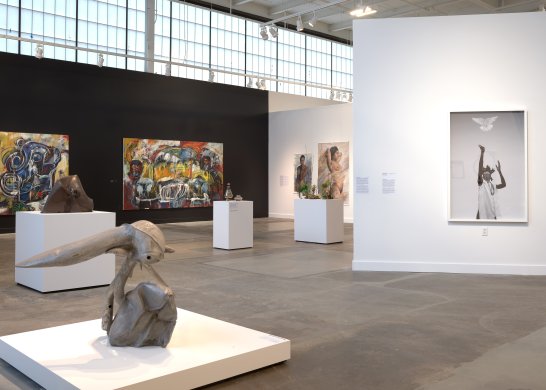 In the background are several paintings on walls and three sculptures on three pedestals. In the foreground are an abstract sculpture on a low pedestal and a black and white photograph.