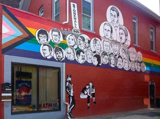 A pantheon of black and white faces over top the Progress Pride Flag is painted on the side of a red brick building