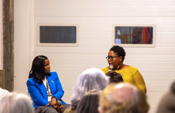 A woman of dark skin tone in a brilliant blue blazer listens as seated next to her a woman of dark skin tone in a deep yellow dress speaks into a microphone