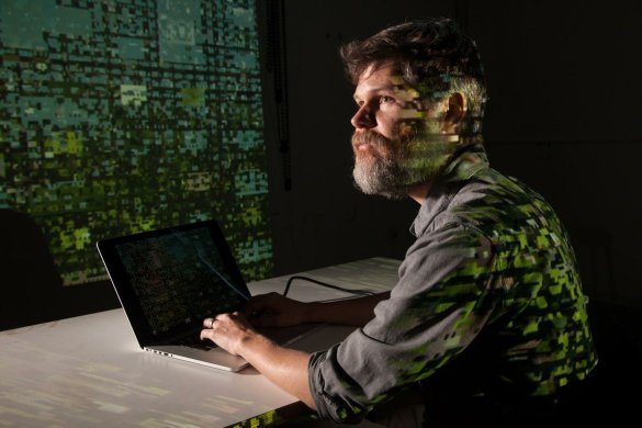 Digital-green light illuminates a man of light skin tone with beard sitting at a laptop