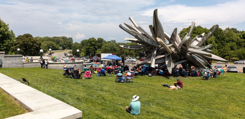 Spectators in lawn chairs sitting around a large outdoor sculpture listening to a band perform