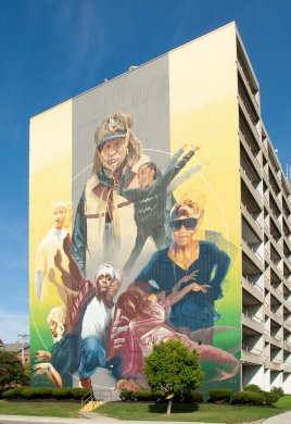 A mural featuring multiple figures on a yellow background on a large concrete apartment building