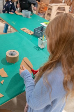 (Photo taken from behind) A young girl with blonde hair and a blue sweater cutting a piece of cardboard over top a green table