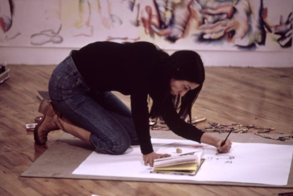 A woman with dark hair kneeling over a sketch pad, drawing on the ground