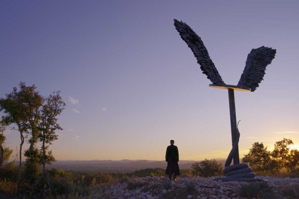 Film still of a person's silhouette next to a large statue of wings outside at sunset