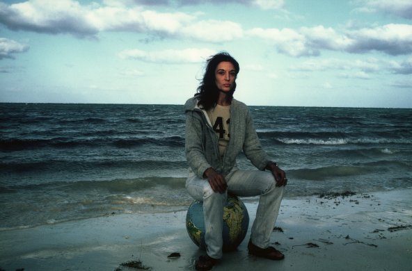 A woman sitting on a globe on a beach wearing jeans and a zip up sweatershirt