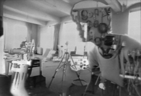 A woman’s face and torso, turned 45 degrees towards the camera, is shown inside (or in front of) a studio space filled with electronic equipment. The woman is half substance and half outline: her shirt is solid, but most her face is a window into another image that shows neatly looped cables hanging on a wall