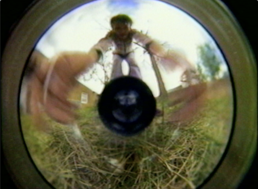 A fisheye lens captures a person reaching towards the camera with grass visible in the background.