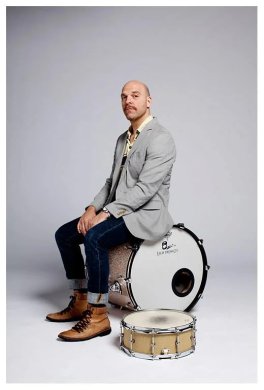 A person sitting on a drum, wearing a suit, in front of a grey background