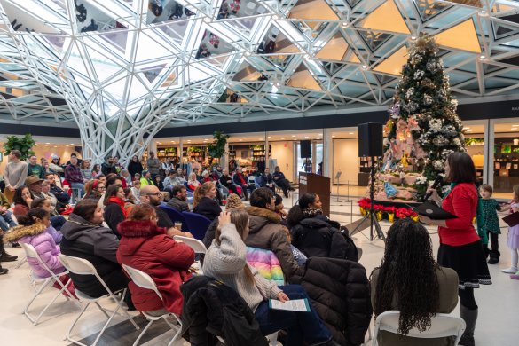 People gathered around a large Christmas tree under a glass mosaic ceiling