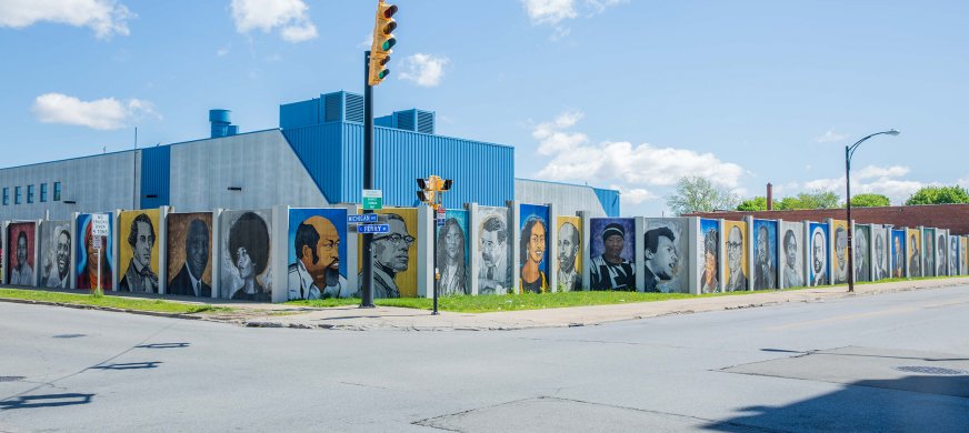 The long concrete wall visible across the center of this image is segmented into twenty-eight vertically oriented rectangles, each filled with the portrait of a local or national civil rights leader. The portraits are painted in four distinct, alternating styles that are more or less photorealistic or stylized and in color or grayscale.