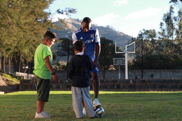 A Black man in a soccer uniform and two kids play with a soccer ball on a soccer field