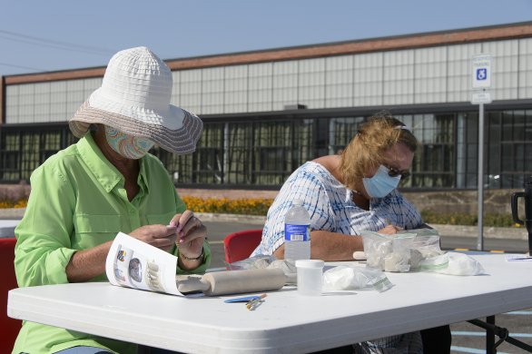 Two white women wearing face masks seated at a table making ceramic beads