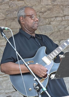 An African American man in a blue shirt playing a blue guitar
