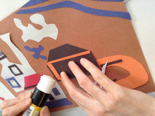 A hand gluing paper shapes to a sheet of brown paper