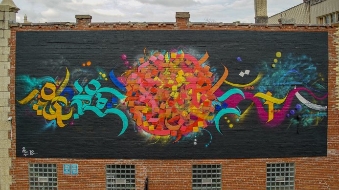 Calligraphic forms in bright shades of orange, turquoise, and magenta pop from a black background painted on a red brick façade. Abstracted from Arabic script, these coalesce in the center into a large, dense circle dominated by pink, red, and yellow forms.